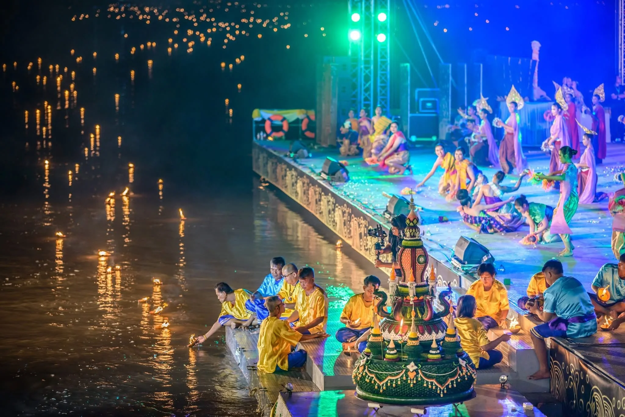 Dancers on a riverside stage beside a river glowing with hundreds of floating lights
