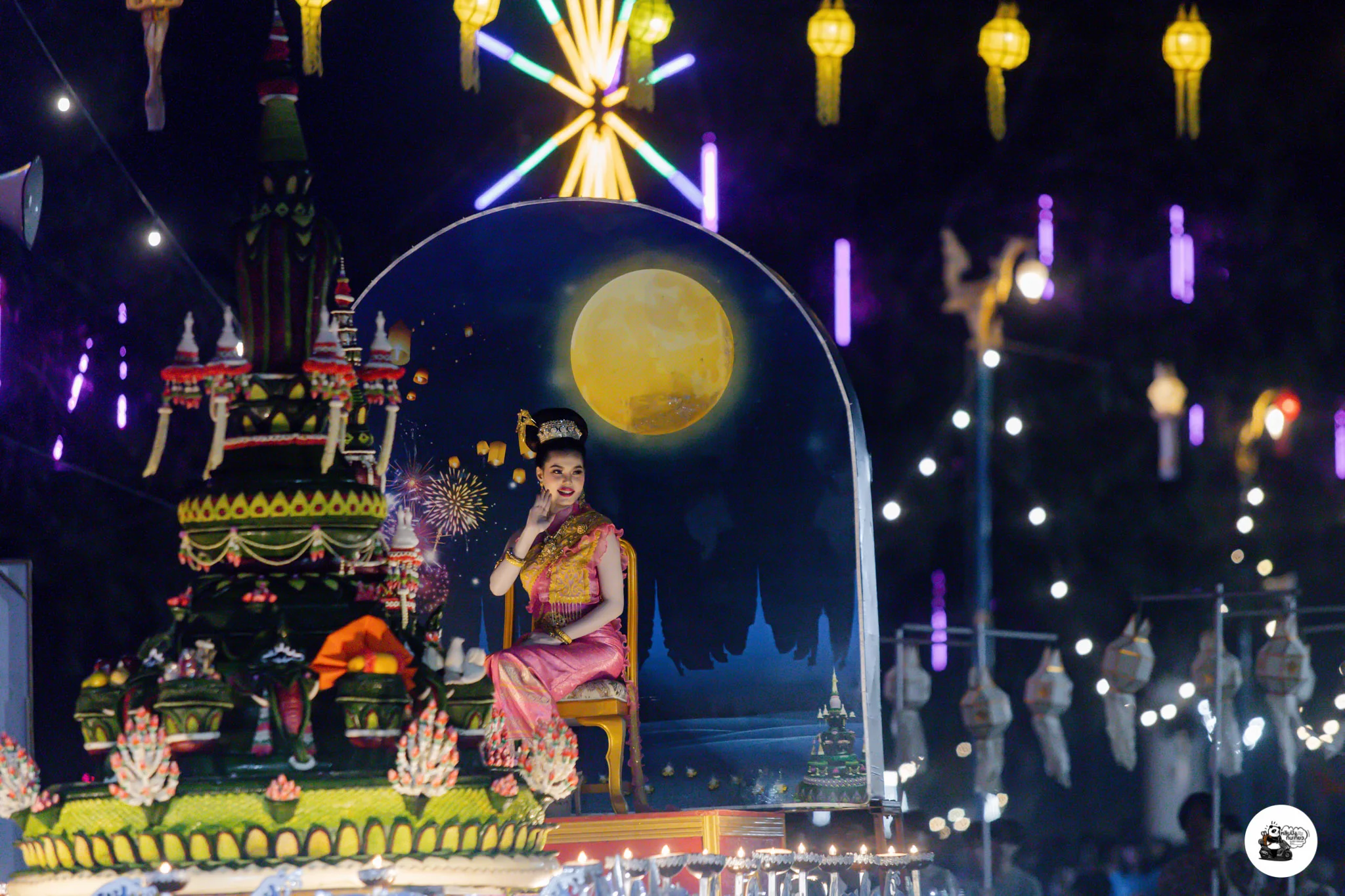 A performer in traditional dress seated on a decorated float in front of a painted full moon backdrop
