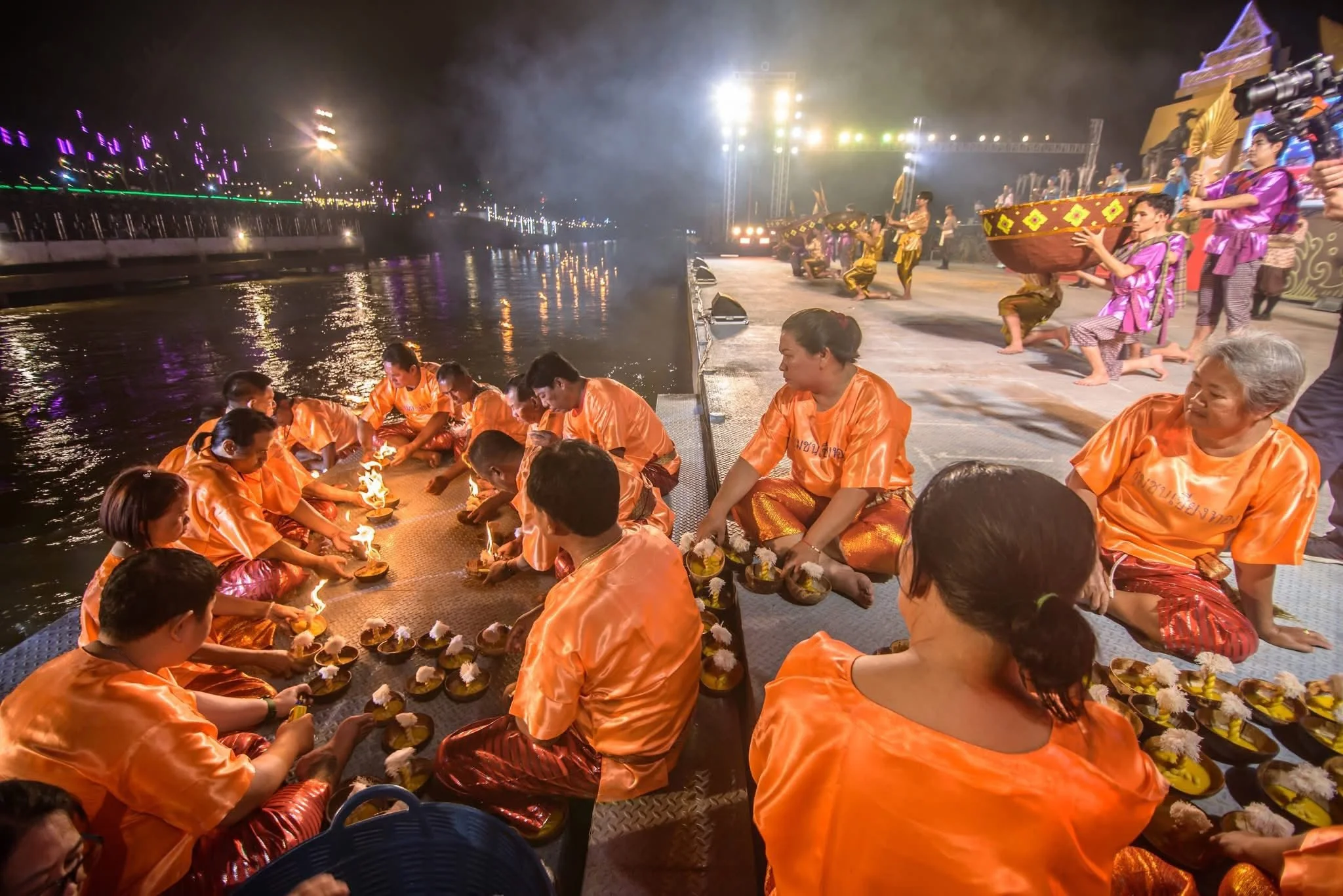 Villagers in orange robes lighting coconut-shell krathongs by the riverbank