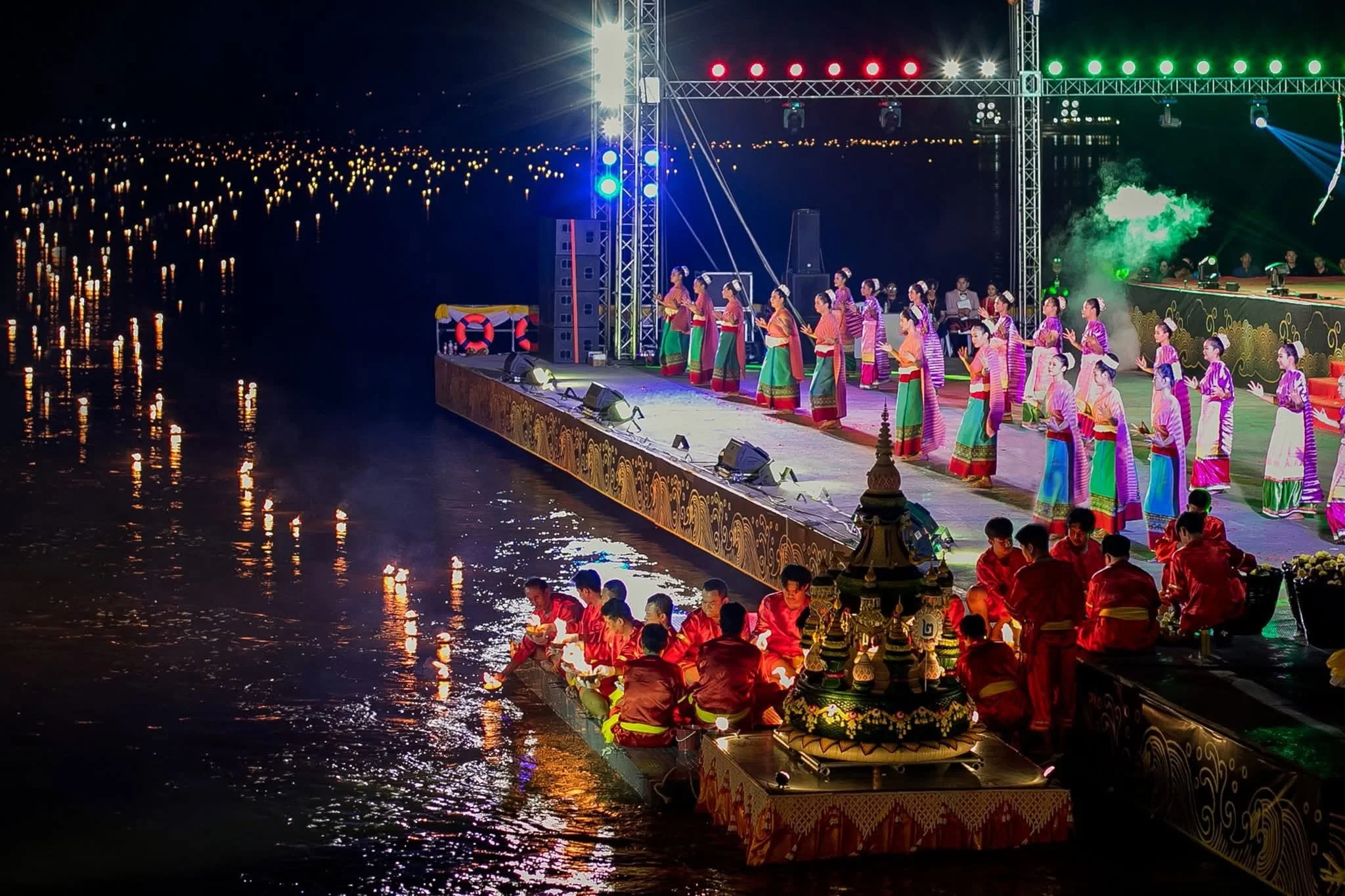 A dance troupe performing on a floating riverside stage, with fireworks behind them