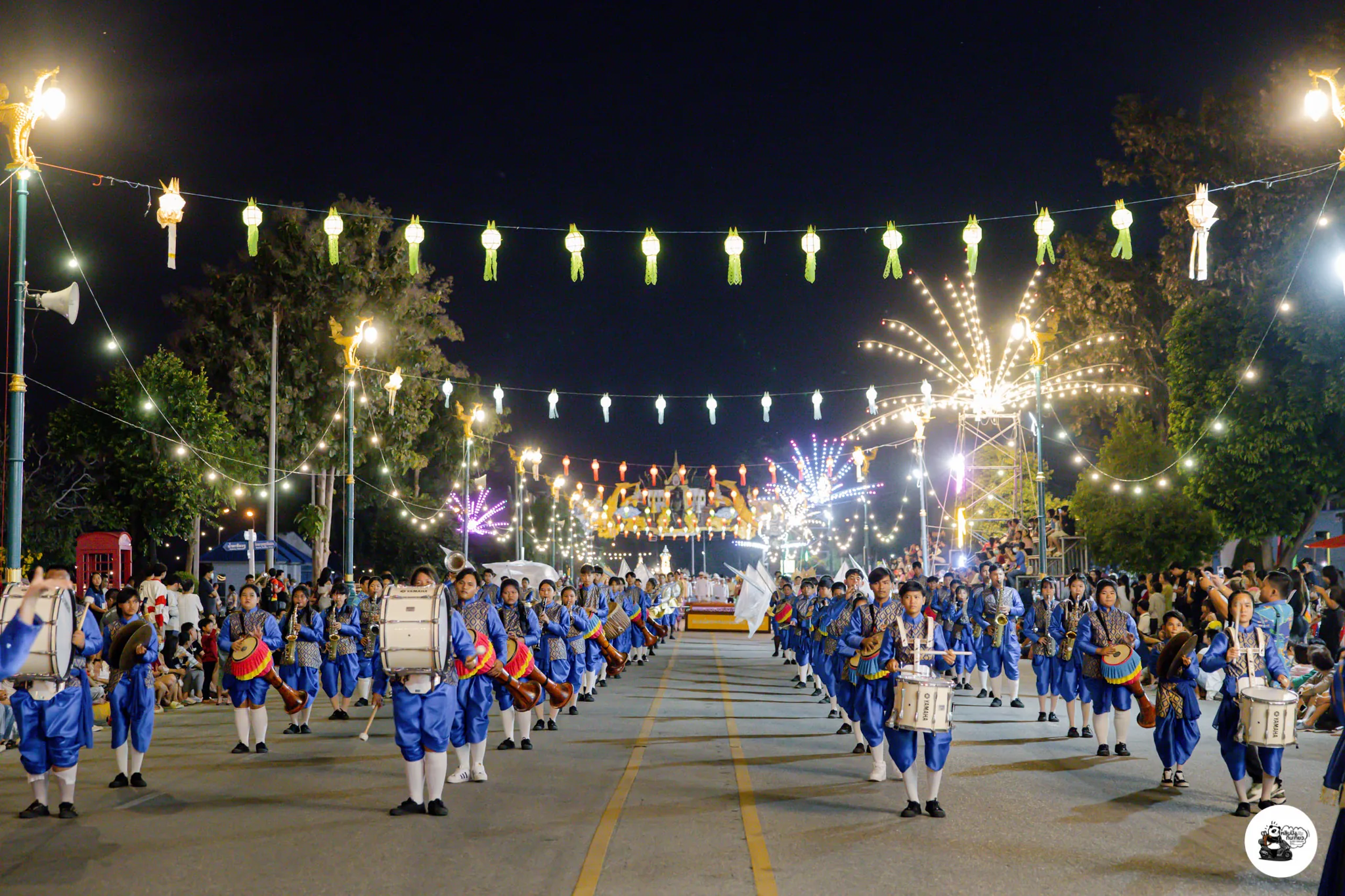 A marching band in traditional dress parading past strings of lanterns at night