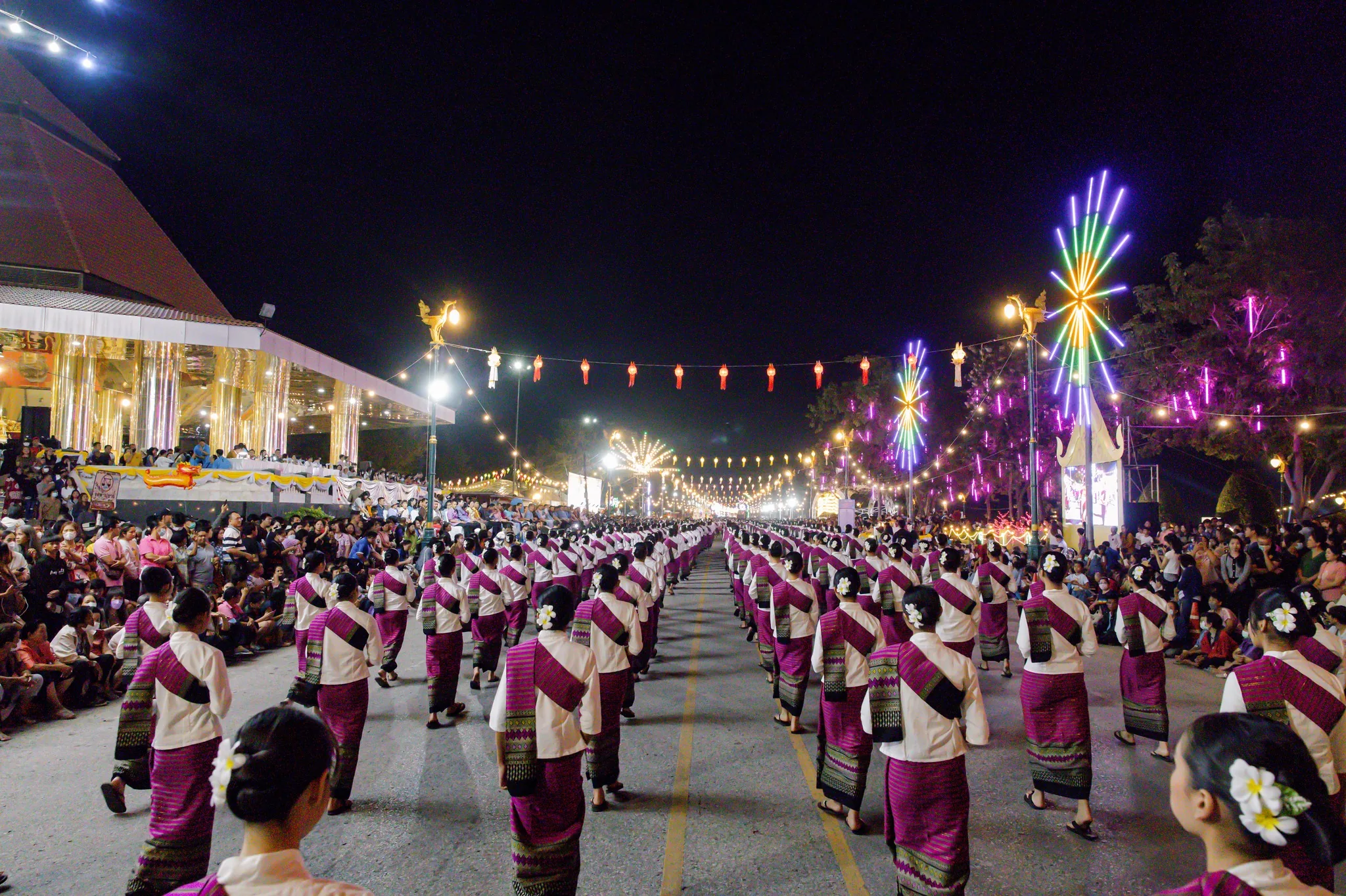 Hundreds of dancers in traditional dress moving in formation down a lit avenue